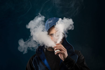 vaping man holding a mod. A cloud of vapor. dark blue background.の写真素材