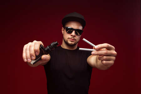Man with cigarettes and e-cigarette on a red background. Choice between dependenciesの写真素材