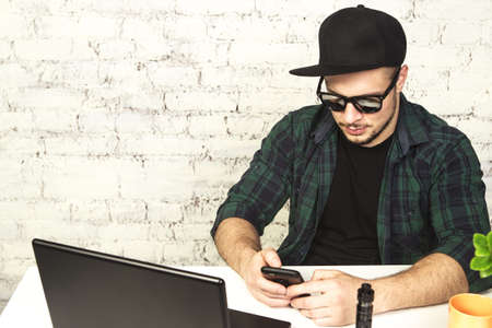 Young man sitting in office with a cup of coffee, vaping and releases a cloud of vapor. Working at laptop and having a little break.の写真素材