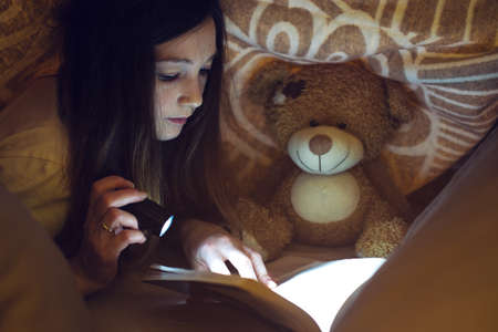 young girl reading a magic book in the dark holding a flashlight in hand under a blanket with a toy teddy bearの写真素材