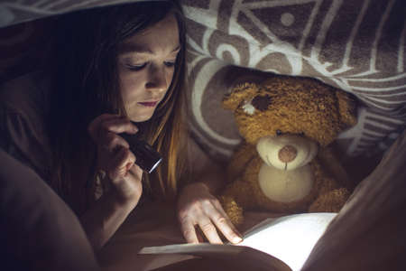 young girl reading a magic book in the dark holding a flashlight in hand under a blanket with a toy teddy bearの写真素材