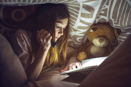 young girl reading a magic book in the dark holding a flashlight in hand under a blanket with a toy teddy bearの写真素材