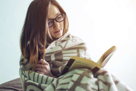 Young woman wrapped in a warm blanket on a winter morning reading an interesting book alone, next to coffee mug and toy bear. The concept of educationの写真素材