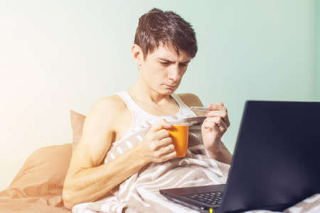 young man sick with a cold lying in bed, unwell with a thermometer wrapped in a blanket and working on the laptopの写真素材