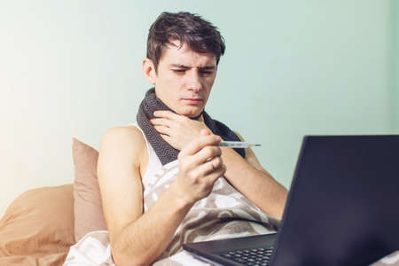 young man sick with a cold lying in bed, unwell with a thermometer wrapped in a blanket and working on the laptopの写真素材