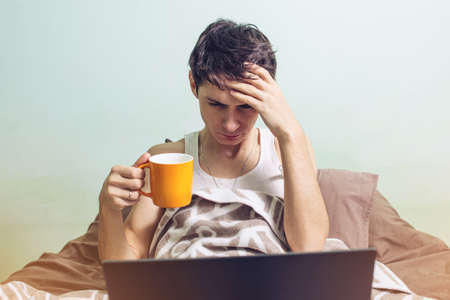 young man sick with a cold lying in bed, unwell with a thermometer wrapped in a blanket and working on the laptopの写真素材