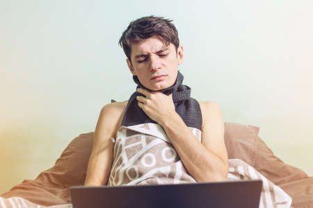 young man sick with a cold lying in bed, unwell with a thermometer wrapped in a blanket and working on the laptopの写真素材