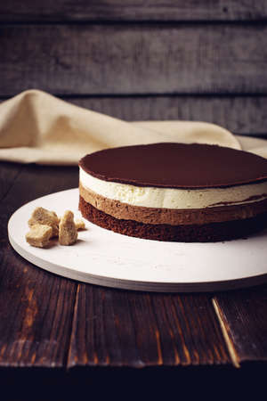 delicious three-layer chocolate cake stands on a circular base, lying next to rushy sugar and tongs on a wooden table and dark background. place for text. food design.の写真素材