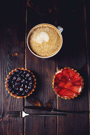 On Berry mini tarts with strawberries and blueberries next a Cup of coffee on a wooden table over dark backgroundの写真素材
