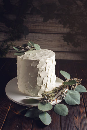 white cream cake decorated with green leaves of the eucalyptus tree on a wooden stand in a rustic...
