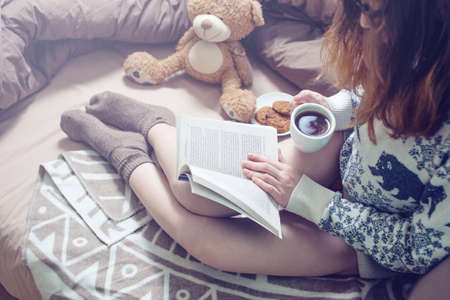 girl reading book in bed in a sweater and warm socks keeping your coffee in hand in the morning. The concept Sunny spring mood.の写真素材