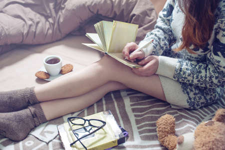 girl writing in a notebook sitting on the bed in a sweater and warm socks keeping your coffee in hand in the morning. The concept of Sunny spring mood.の写真素材