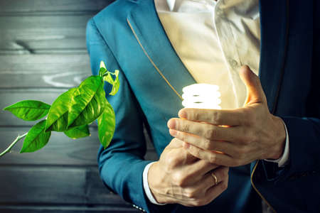 Man businessman in suit holding a glowing light near the green sprout of a young tree, plant on dark wooden background. Concept of environmental energy, ecology of planet, careful use of electricityの写真素材