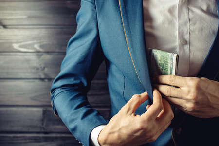 Man businessman, member or officer in a suit puts a bribe in the form of hundred dollar bills in his pocket. the concept of corruption and bribery among high-ranking officialsの写真素材