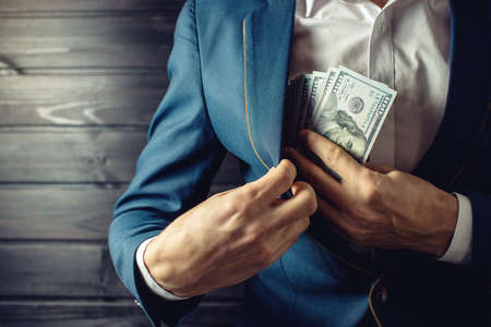 Man businessman, member or officer in a suit puts a bribe in the form of hundred dollar bills in his pocket. the concept of corruption and bribery among high-ranking officialsの写真素材
