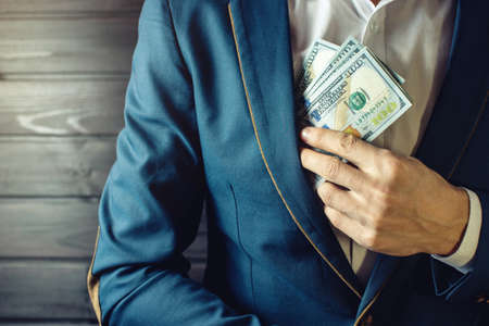 Man businessman, member or officer in a suit puts a bribe in the form of hundred dollar bills in his pocket. the concept of corruption and bribery among high-ranking officialsの写真素材