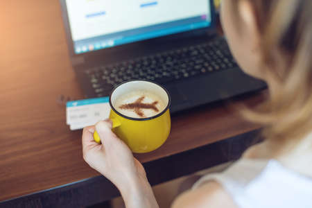 Woman holds in her hands a Cup of coffee with the plane of the cinnamon working at a laptop and to reserve or buy tickets online. Travel conceptの写真素材