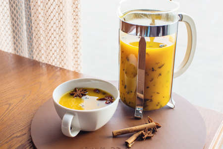 Cup and glass teapot with spicy hot fruit tea with orange and mango next to an open book on table in cafeの写真素材