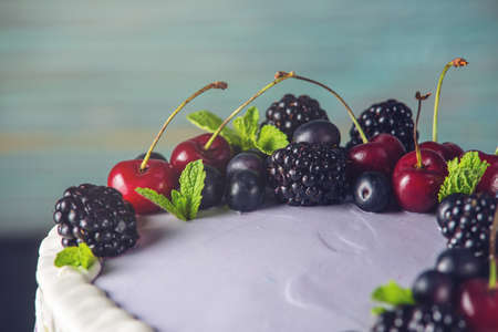 Beautiful purple cake with streaks of white cream and decorated with berries BlackBerry, blueberry and cherry. Over blue wooden table. Dark rustic style.の写真素材