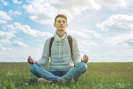 Man traveler with backpack sits on meadow with green grass under blue sky with clouds in the Lotus...