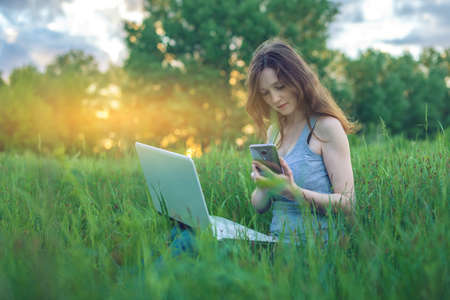 Woman sitting on a green meadow with trees on the background of sunset with clouds. Working or studying on laptop wireless and looking at the phone. The concept of education with natureの写真素材