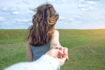 Follow me, Attractive brunette girl holding the hand of the leads in a clean green field, steppe with clouds. The concept of the journey to the meeting of the natureの写真素材