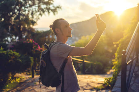 Man traveler with backpack makes a photo on his smartphone outdoors with mountains in the background. The concept of connection and communication in the journey.の写真素材