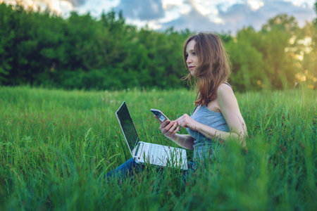 Woman sitting on a green meadow with trees on the background of sunset with clouds. Working or...