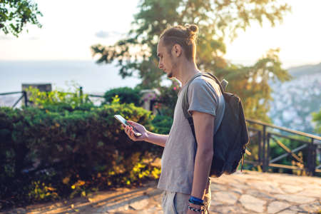 Man traveler with backpack looking at his smartphone on the background of nature and the mountains. The concept of connection and communication in the journey.の写真素材