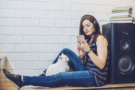 Woman in headphones listening to an audiobook on a phone sitting with cat on wall background. Concept of technology educationの写真素材