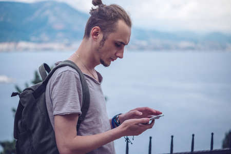 Man traveler with backpack looking at his smartphone on the background of nature and the mountains. The concept of connection and communication in the journey.の写真素材