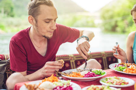 Young man eats vegetables and fish outdoors in a cozy restaurant on the water during travel, discover new kitchenの写真素材