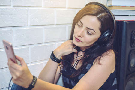 Woman in headphones listening to an audiobook on a phone sitting sitting on wall background. the concept of technology educationの写真素材