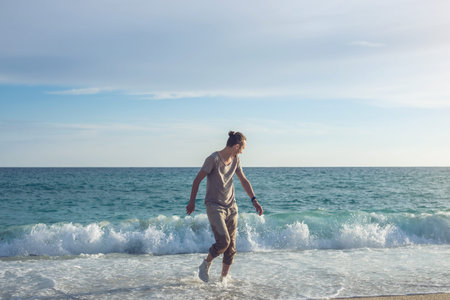 Young happy man walking in wet sneakers from the sea on the shore and running away from waves. The concept of travel and freedomの写真素材