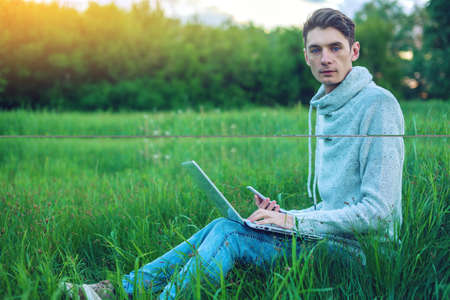 Young man sitting on a green meadow with laptop and using the phone on the background of blue cloudy sky at sunset. The concept of education and business on natureの写真素材