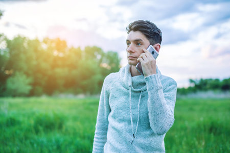 Young man businessman talking at mobile phone outside on a grass field at sunset