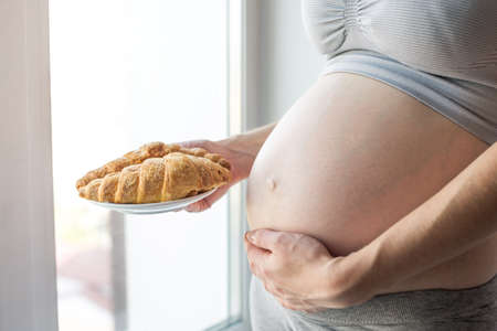 A pregnant woman with belly holding a plate of croissants. The concept weight control and an unhealthy diet during pregnancyの写真素材