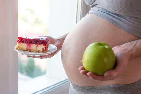 A pregnant woman belly holding a plate with junk and healthy food. The concept for weight control and choice of diet during pregnancyの写真素材