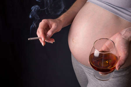 A pregnant woman with a big belly holding a glass of whiskey and a cigarette in hands on dark background. The concept of Smoking and bad habits during pregnancyの写真素材