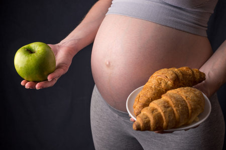 A pregnant woman belly holding a plate with junk and healthy food. The concept for weight control...