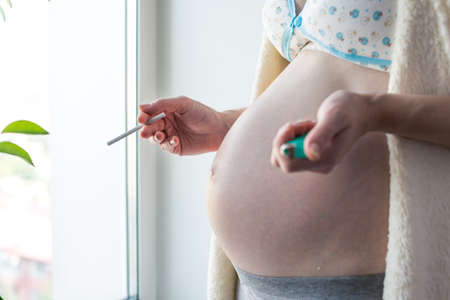 A pregnant woman with a big belly, smokes by the window holding a coffee and a cigarette. The concept of Smoking and bad habits during pregnancyの写真素材