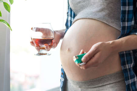 A pregnant woman with a big belly by the window holding a glass of whiskey and a cigarette. The concept Smoking, alcohol and bad habits during pregnancyの写真素材