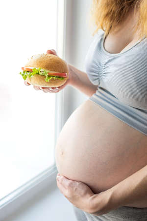 A pregnant woman with belly by the window holding a burger. The concept of fast food and unhealthy eating during pregnancyの写真素材