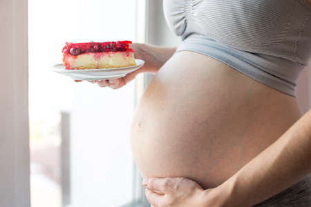 A pregnant woman with belly holding a piece of cheesecake. The concept weight control and an unhealthy diet during pregnancyの写真素材