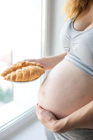 A pregnant woman with belly holding a plate of croissants. The concept weight control and an unhealthy diet during pregnancyの写真素材
