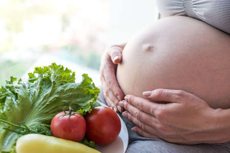 A pregnant woman with belly holding a plate with vegetables in hands. The concept for weight control and healthy eating during pregnancyの写真素材