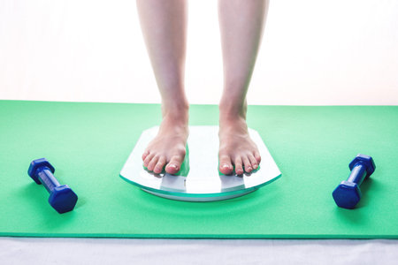 Female feet standing on electronic scales for weight control, dumbbells and measuring tape. The...