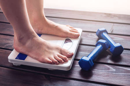 Female feet standing on mechanical scales for weight control, dumbbells and measuring tape on wooden background. The concept of sports training, slimming and weight lossの写真素材