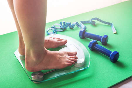 Female feet standing on electronic scales for weight control, dumbbells and measuring tape. The concept of sports training, slimming and weight lossの写真素材