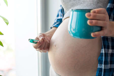A pregnant woman with a big belly, smokes by the window holding a coffee and a cigarette. The concept of Smoking and bad habits during pregnancyの写真素材